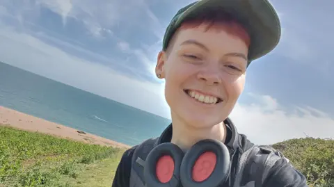 Hat A woman stood on a hill with a beach and the sea behind her. She is smiling and has on a baseball cap and large headphones around her neck.