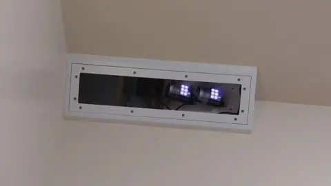 The Oxevision technology - which is an infrared camera - placed near the ceiling in the corner of a bedroom. It is in a white rectangle box and small white lights can be seen at one end.