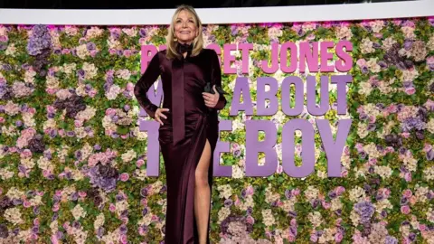 Helen Fielding avec une robe bordeaux devant un mur floral.