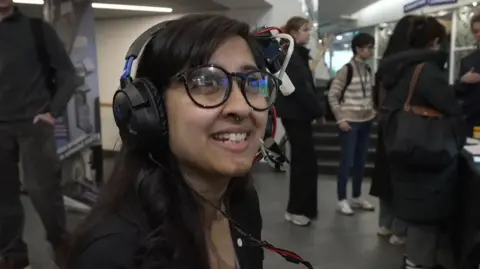Une femme portant un casque expérimental sourit pendant qu'elle regarde hors caméra.