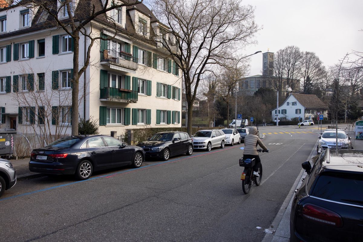 Une rue à Wollishofen, Zurich, avec des voitures garées et un cycliste. Sujet : suppression de places de stationnement pour la construction de la Velovorzugsroute.