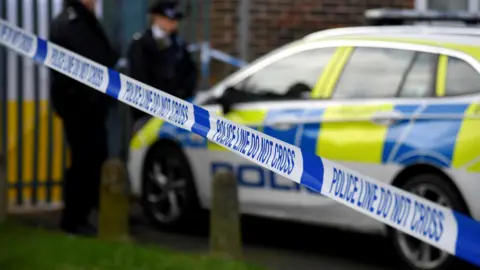 Deux policiers, flous, se tiennent près d'une voiture de police derrière un ruban de police alors qu'ils enquêtent sur une scène de meurtre à l'arme blanche à Croydon
