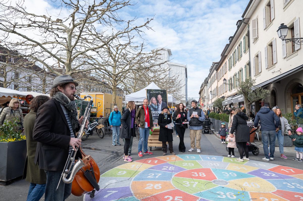 Place du Marché à Carouge avec des stands pour les élections municipales 2025, des personnes se rassemblant autour.