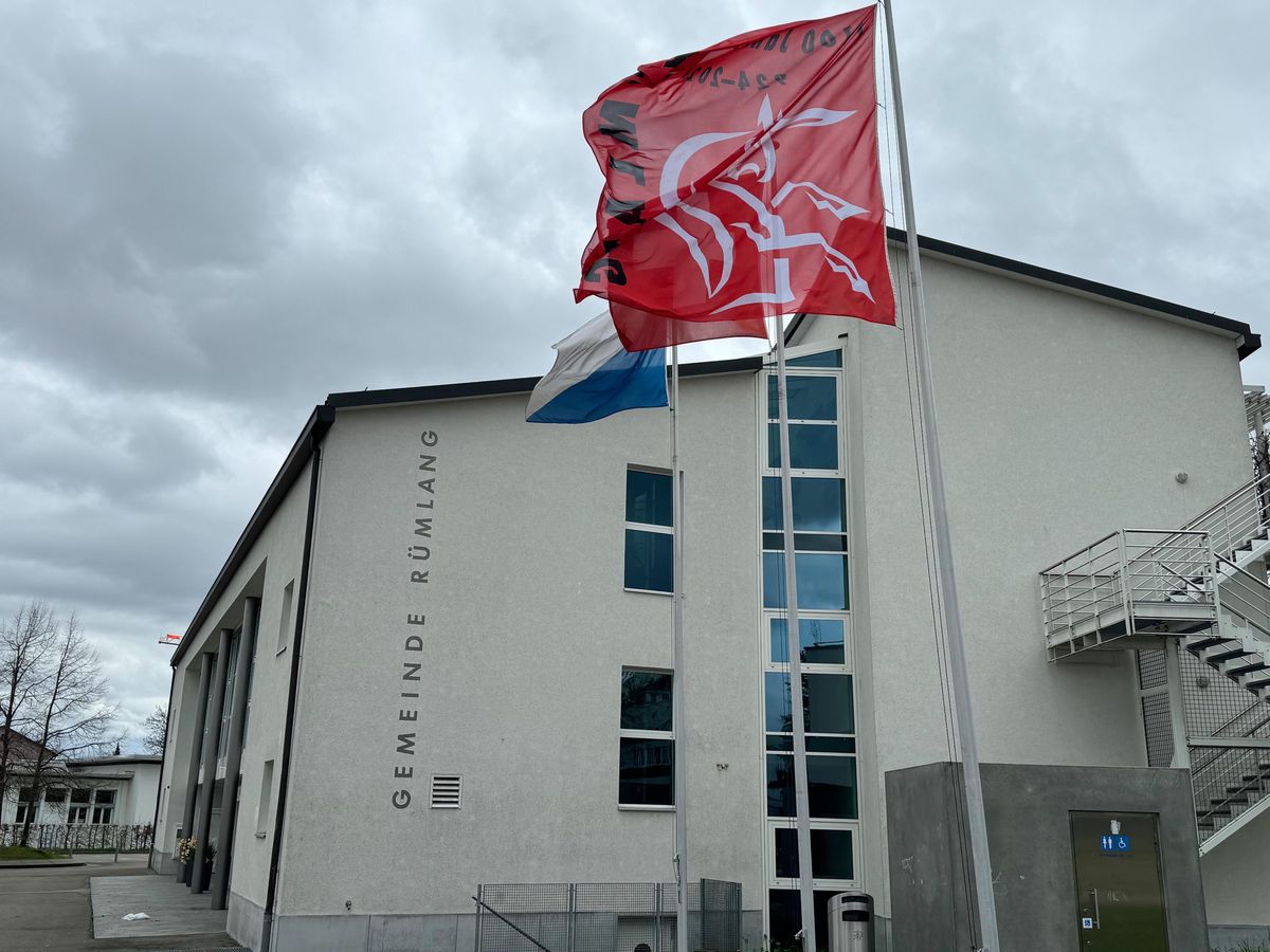 Gemeindehaus Rümlang avec des drapeaux flottant sous un ciel nuageux.