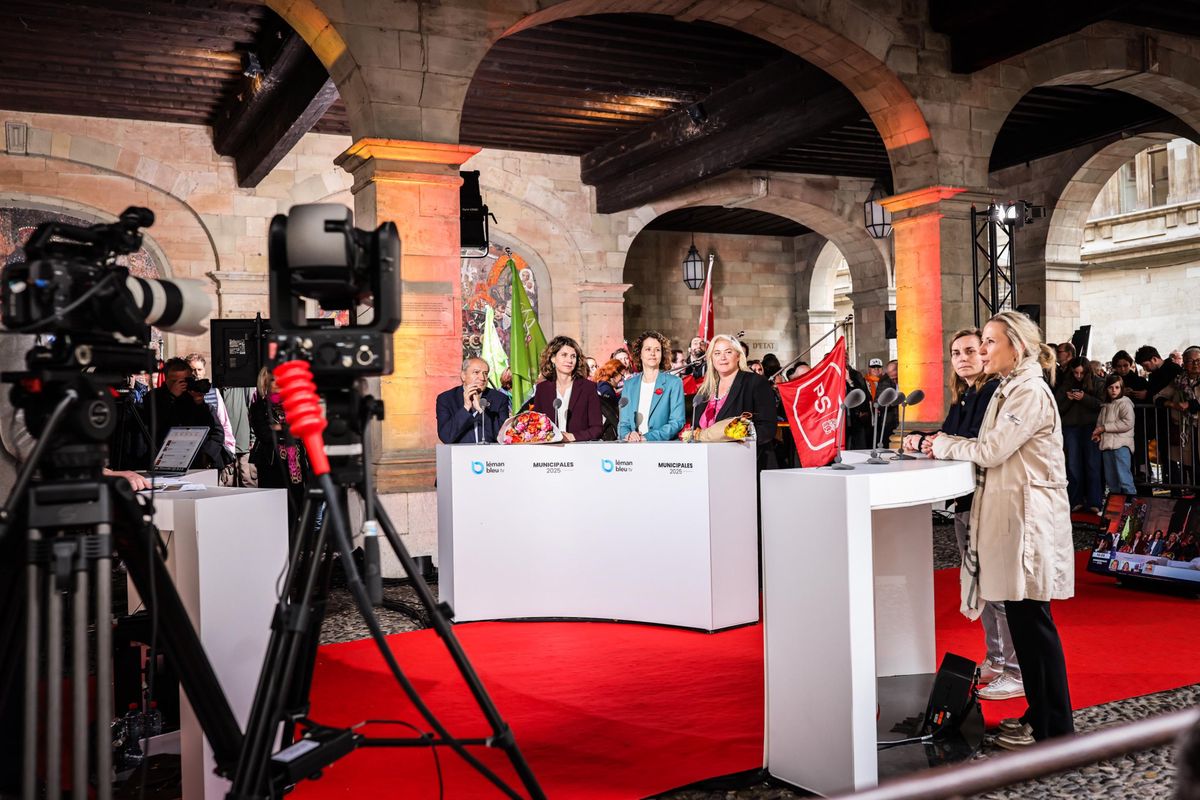 Un événement médiatique se déroule sous un portique avec des participants debout sur un tapis rouge, un appareil photo en premier plan.