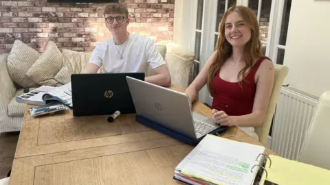 Ben et Emma à la table de la cuisine avec livres et ordinateurs, souriant à la caméra