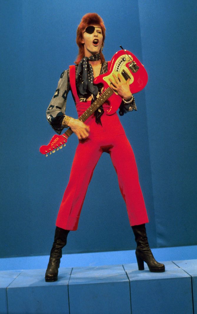 Portrait de David Bowie en 1976, interprétant ’Rebel Rebel’ aux studios TopPop TV en Hollande, vêtu d’un costume rouge et tenant une guitare.