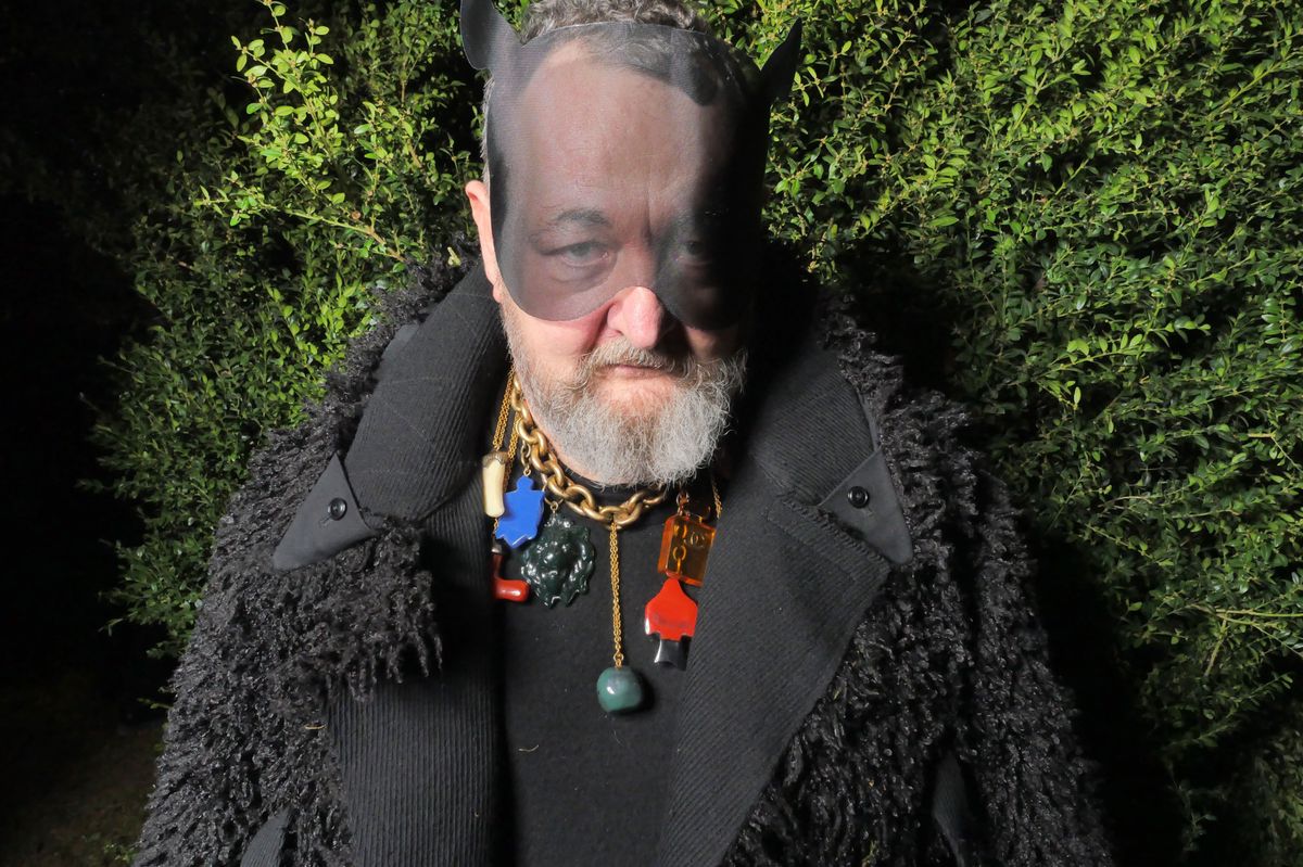 Michel Gaubert portant un masque à cornes noires et un manteau en fourrure, debout devant des buissons.