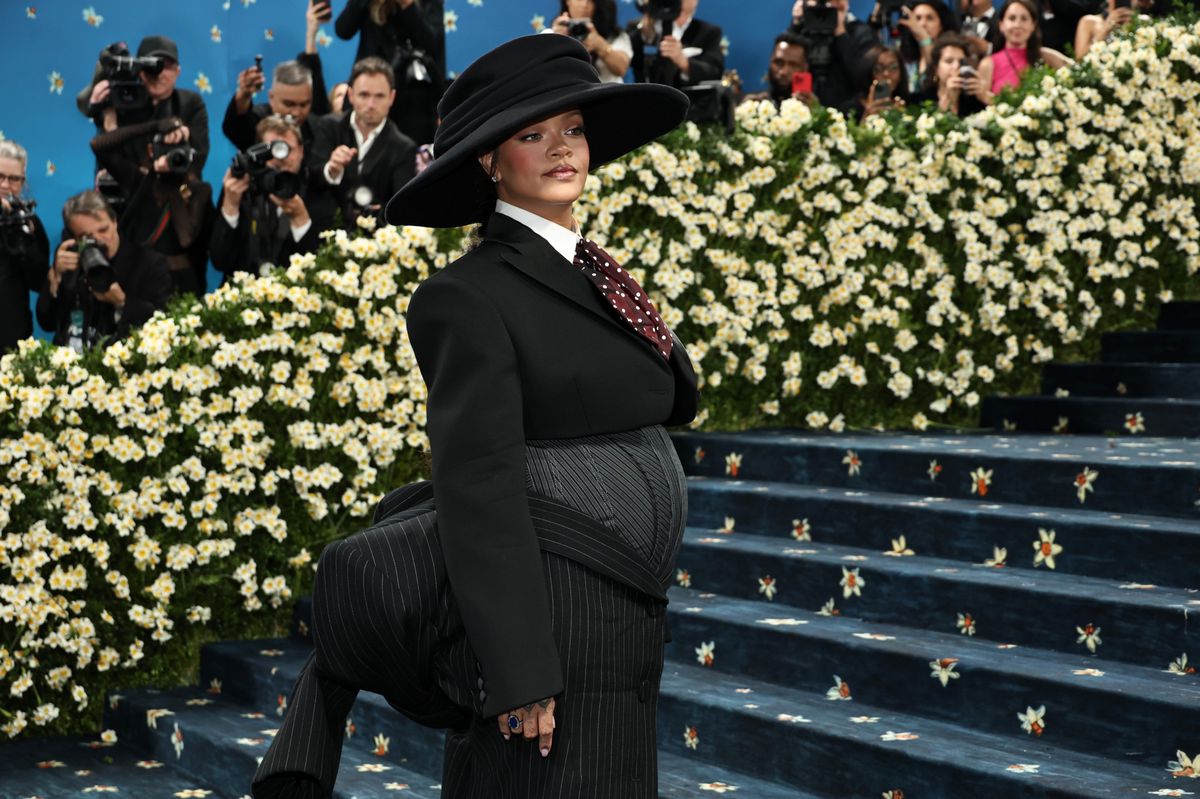 Rihanna au Met Gala 2025, portant une tenue de style tailleur noir avec un grand chapeau, devant des escaliers fleuris au Metropolitan Museum of Art, à New York.