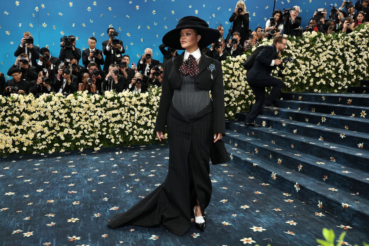 Rihanna au Met Gala 2025, portant une tenue noire élégante et un chapeau large, entourée de photographes et de fleurs blanches au Musée d’art métropolitain de New York.