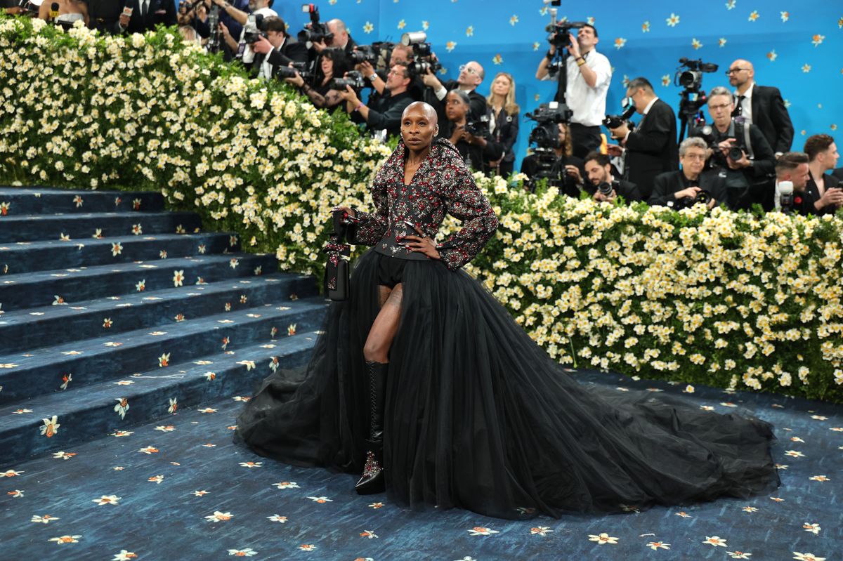 Cynthia Erivo portant une robe noire à longue traîne au Met Gala 2025, entourée de fleurs blanches et de photographes.
