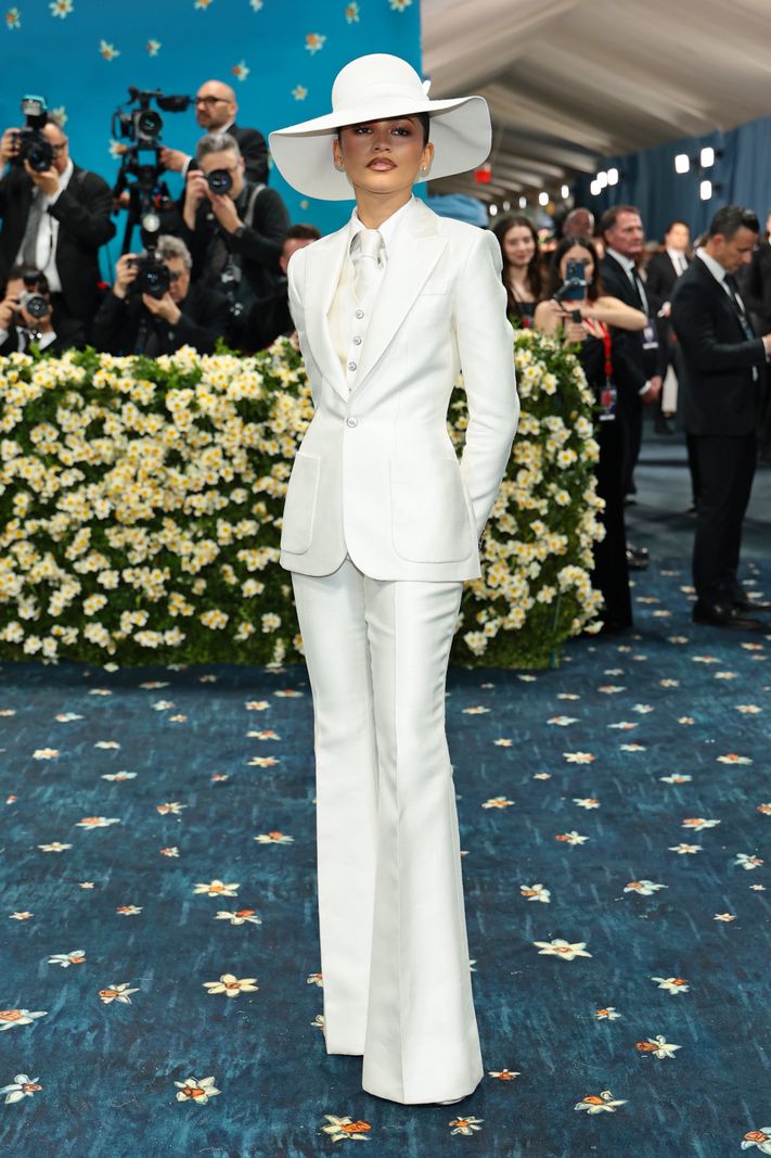 Zendaya au Met Gala 2025, portant un costume blanc élégant et un chapeau à large bord, posant sur un tapis bleu fleuri, entourée de photographes.