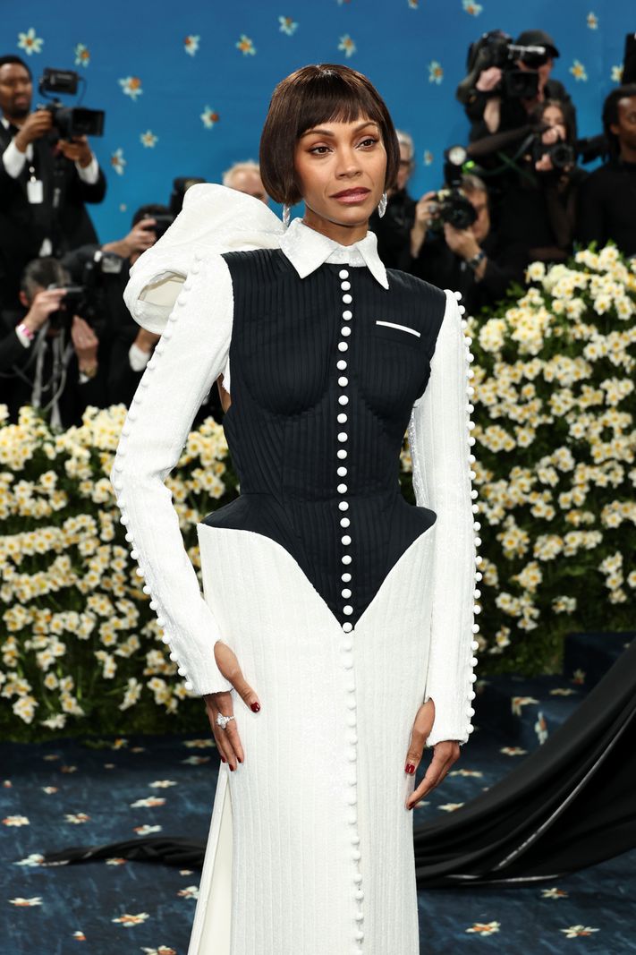 Zoe Saldana au Met Gala 2025, portant une robe noir et blanc à boutons, entourée de fleurs blanches et de photographes.
