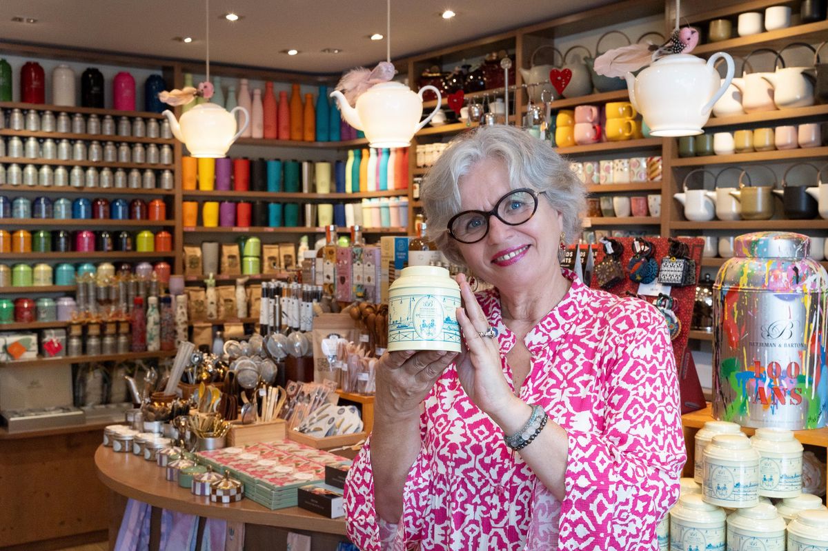 Femme âgée souriante, tenant un pot de thé, dans une boutique remplie de théières et tasses colorées exposées sur des étagères.