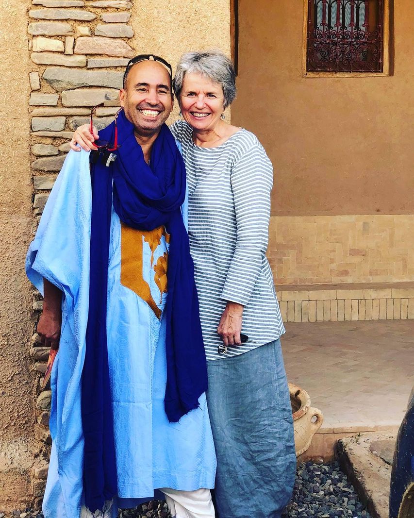 Deux personnes souriantes posant ensemble devant un bâtiment en pierre, l’une portant un vêtement bleu traditionnel.