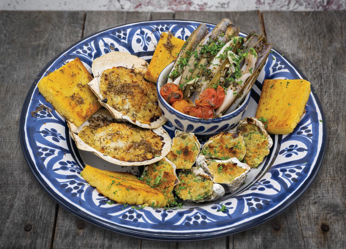 Assiette de fruits de mer variés, comprenant des palourdes gratinées, des huîtres et des couteaux, accompagnée de légumes grillés et de polenta sur un plateau décoratif bleu et blanc.