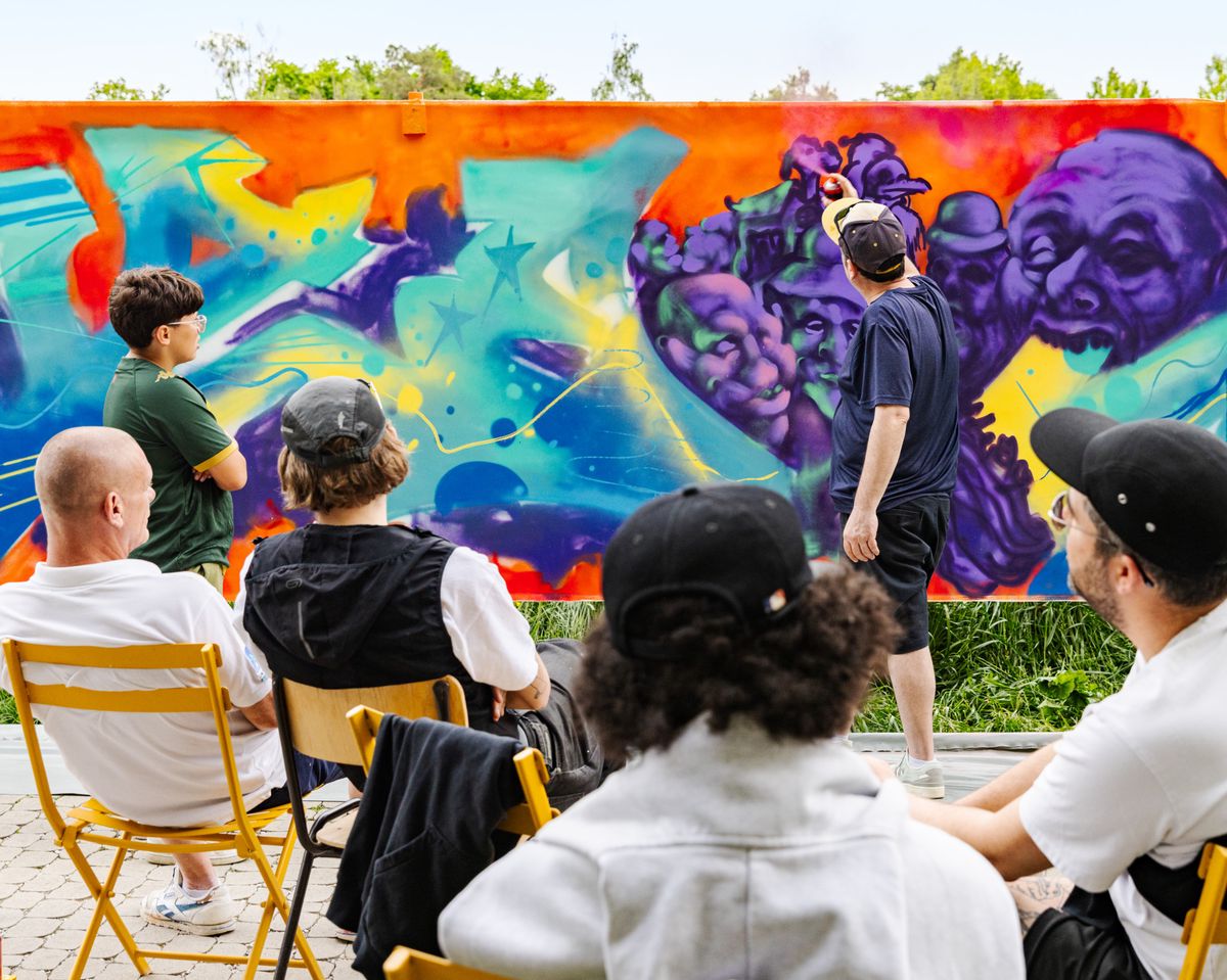 Un groupe de personnes assises regardant un artiste peindre une fresque murale colorée en plein air.