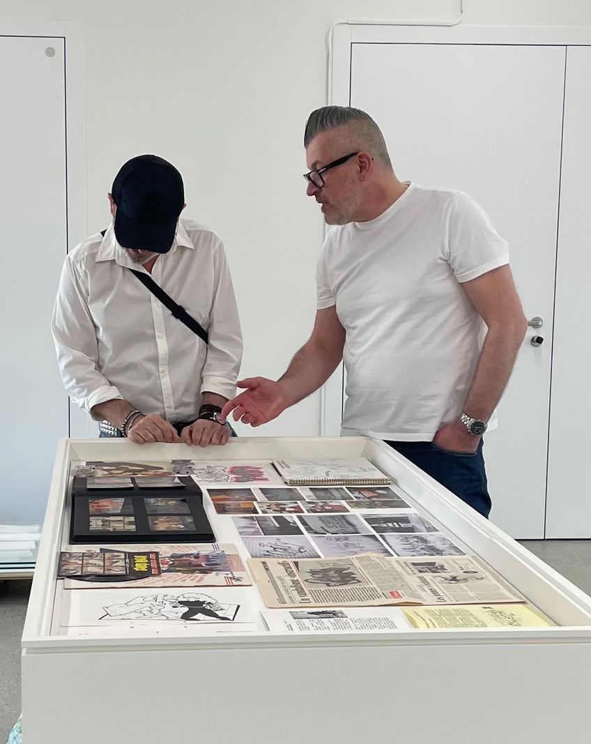 Deux hommes discutent devant une vitrine exposant des documents et des images dans une galerie.