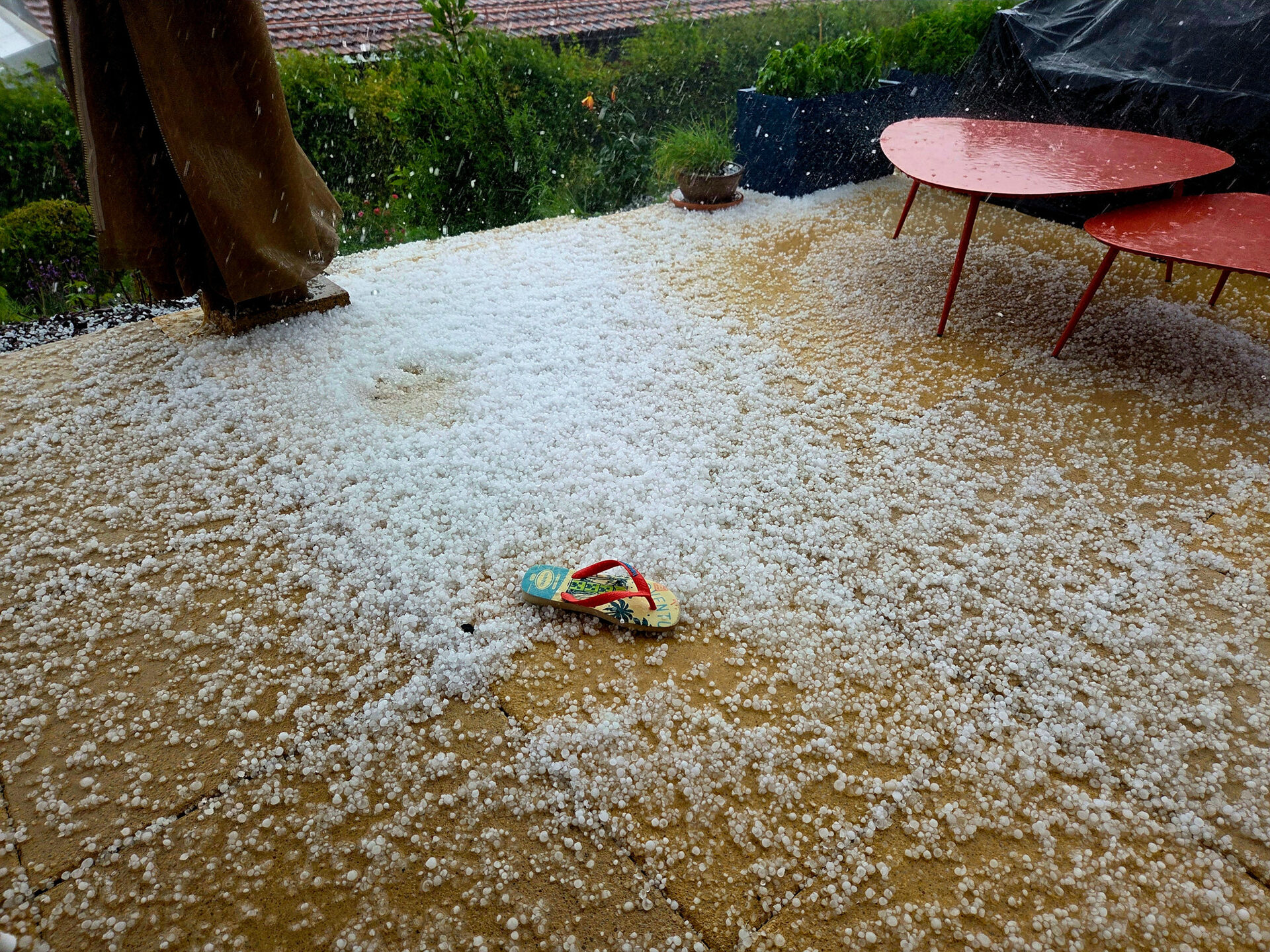 Une violente tempête de grêle couvre une terrasse extérieure avec un mélange de grêle et de neige. Une unique sandale est laissée par terre, entourée par de la grêle, tandis que deux tables rouges sont visibles sur le côté droit.