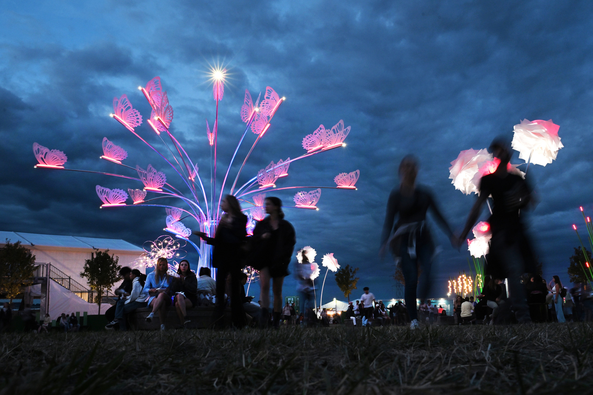 Ambiance nocturne au Paléo Festival 2025 à Nyon, avec des sculptures lumineuses en forme de fleurs et de papillons, ciel nuageux en arrière-plan.