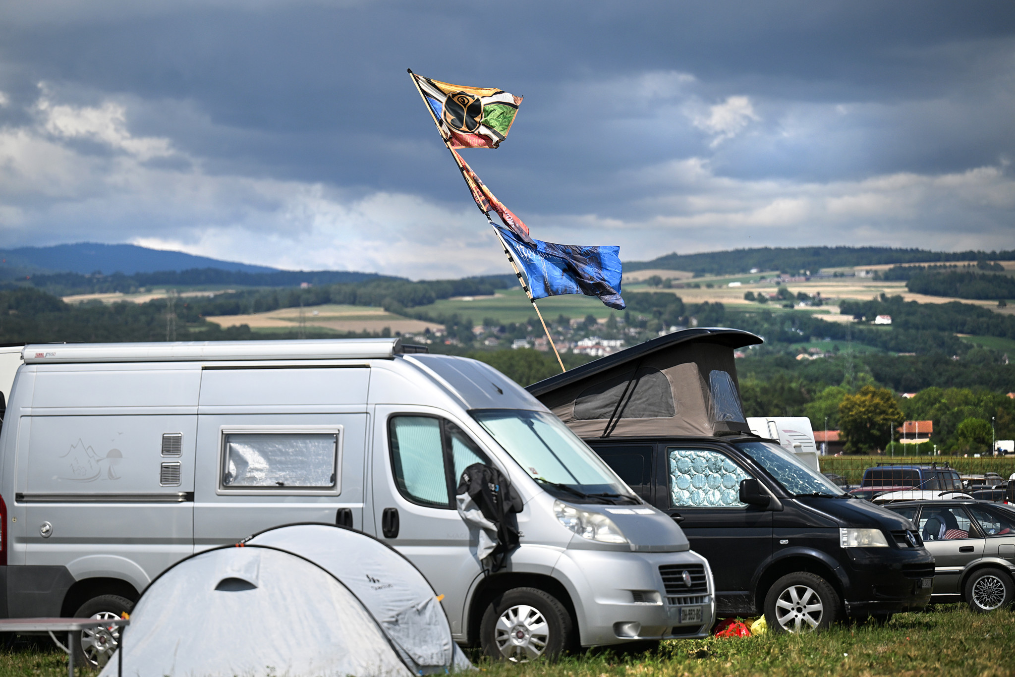 Vans de camping et tentes au camping de Paléo, à Nyon, sous un ciel nuageux, avec des drapeaux colorés flottants.