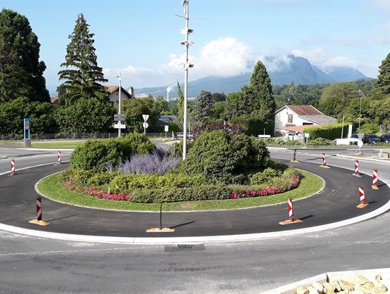 Un rond-point paysager bordé de barrières de chantier, entouré de verdure avec une montagne en arrière-plan.