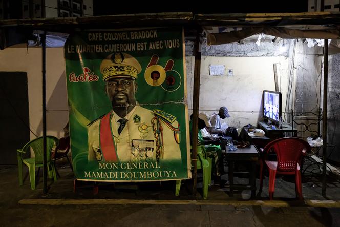 Habitants regardent télévision devant affiche Doumbouya